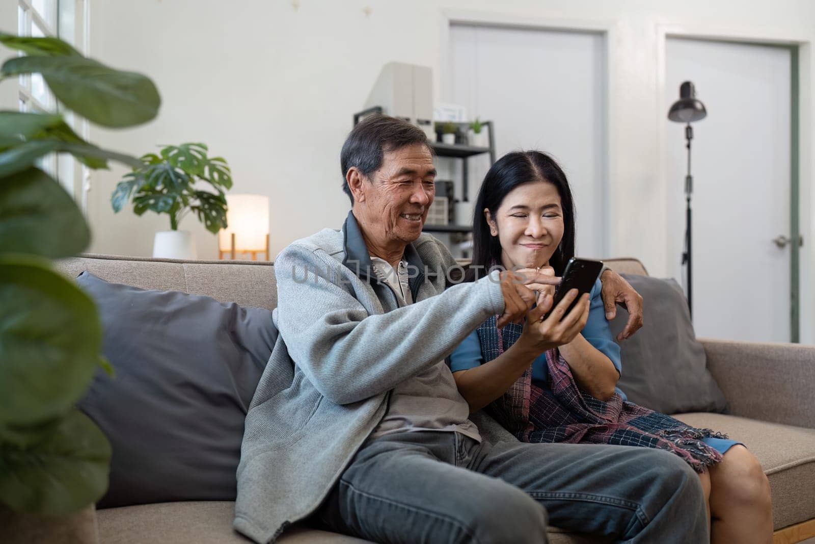 Happy married retirement couple looking at mobile using smartphone mobile technology device together, relaxing on couch doing online shopping.