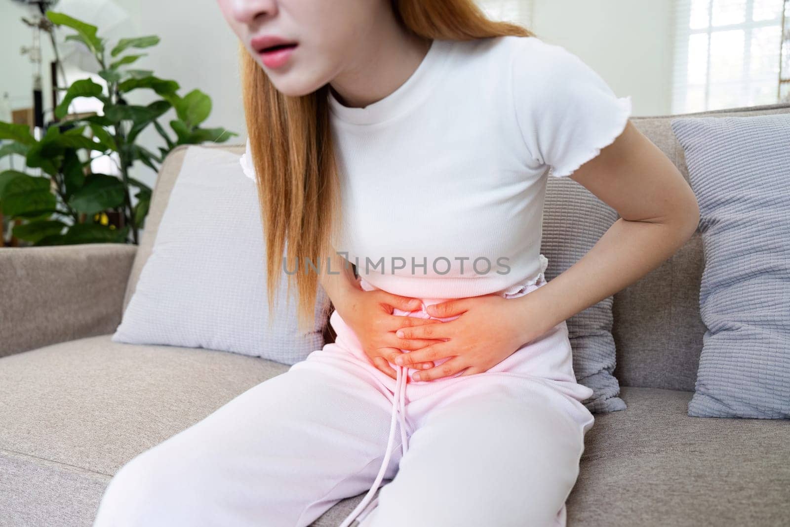 A woman is sitting on a couch with a stomach ache. She is holding her stomach and she is in pain