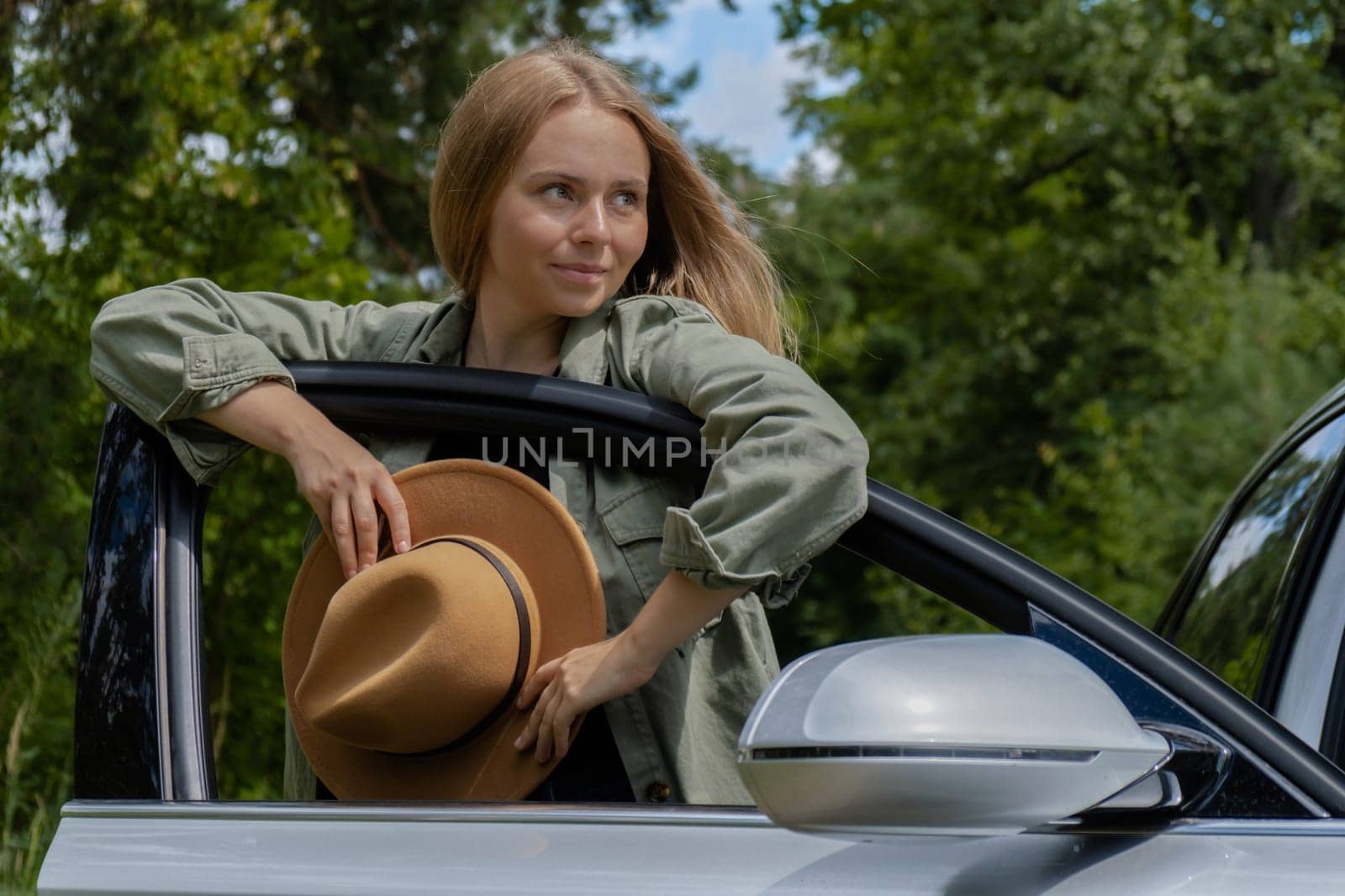 Smiling young woman in hat stoped on road to make a break in driving car. Local solo travel on weekends concept. Exited woman explore freedom outdoors in forest. Unity with nature lifestyle, rest recharge relaxation