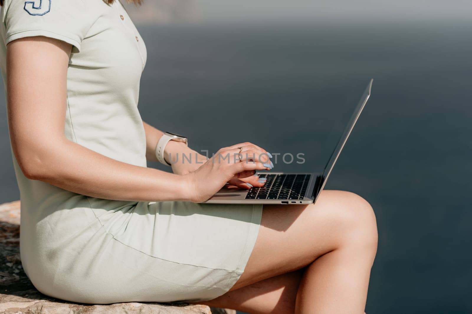 Digital nomad, woman in the hat, a business woman with a laptop sits on the rocks by the sea during sunset, makes a business transaction online from a distance. Freelance, remote work on vacation.