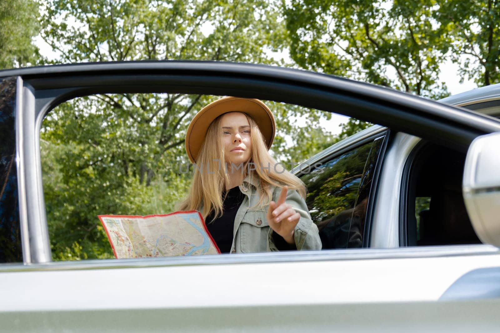 Blonde woman in hat staying next to car door checks the route on map to get to the destination. Young tourist explore local travel making candid real moments. True emotions expressions of getting away and refresh relax by anna_stasiia