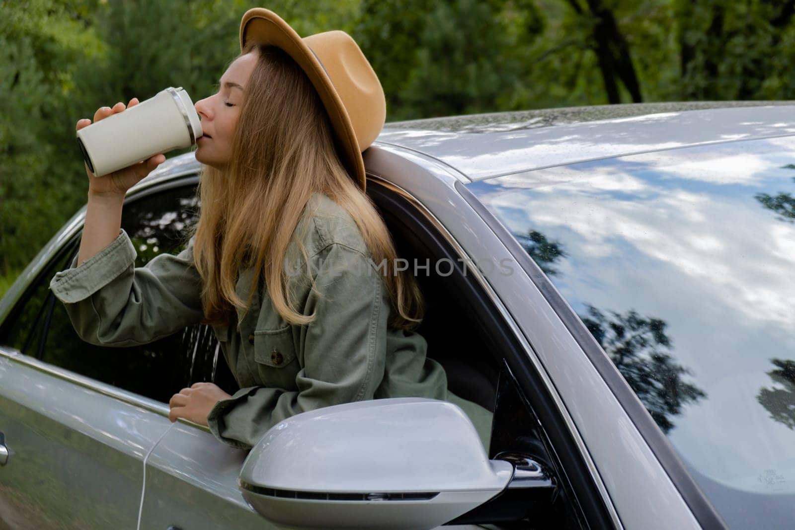Smiling young woman looking from car window and drinking coffee or tea from reusable thermos cup. Local solo travel on weekends concept. Exited woman explore freedom outdoors in forest. Unity with nature lifestyle, rest recharge relaxation