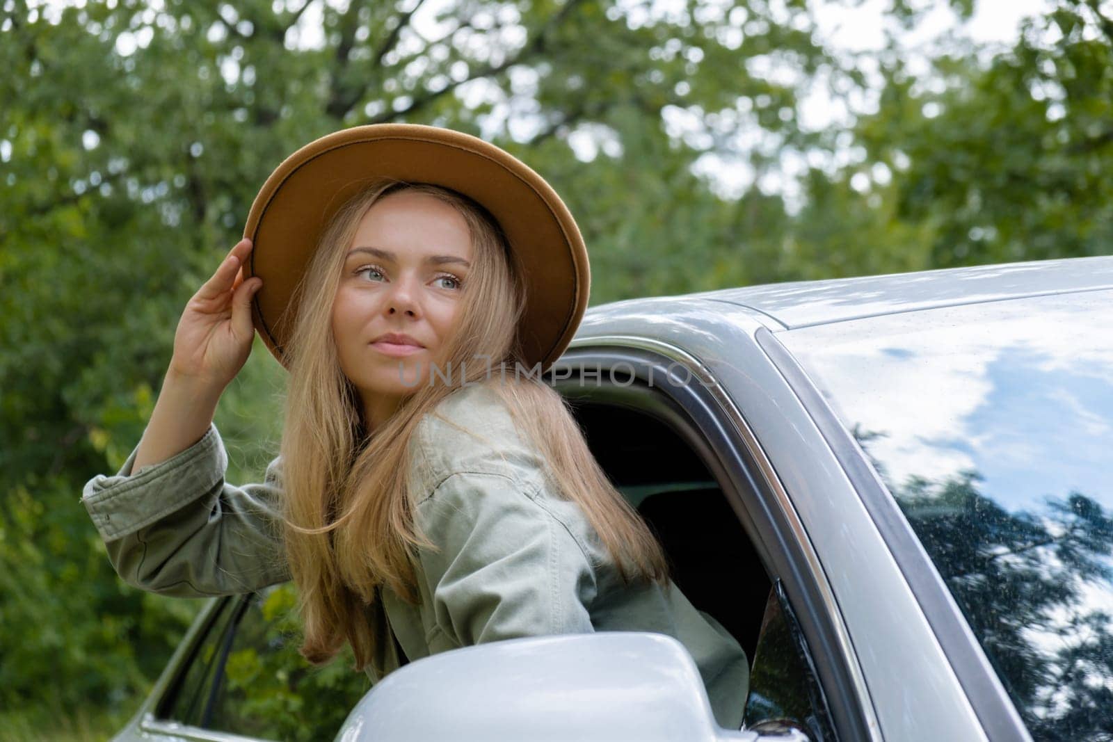 Smiling young woman in hat looking from car window. Local solo travel on weekends concept. Exited woman explore freedom outdoors in forest. Unity with nature lifestyle, rest recharge relaxation