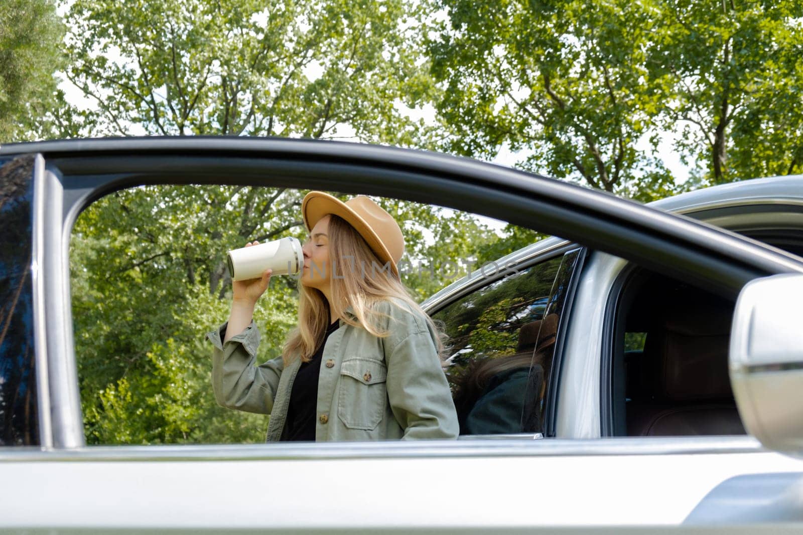 Blonde woman stoped on road next to car and drink coffee or tea from reusable mug. Refuse reuse recycle zero waste concept. Young tourist explore local travel making candid real moments. Responsible traveling reduce carbon footprint by anna_stasiia