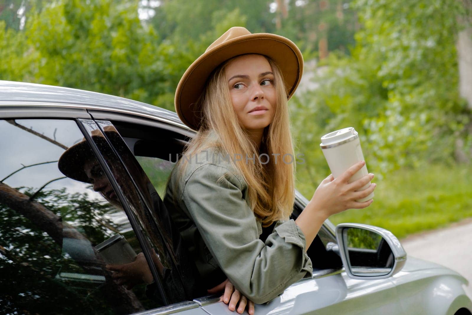 Blonde woman sticking head out of windshield car and drink coffee or tea from reusable mug. Young tourist explore local travel making candid real moments. Refuse reuse recycle zero waste concept. Responsible traveling reduce carbon footprint by anna_stasiia