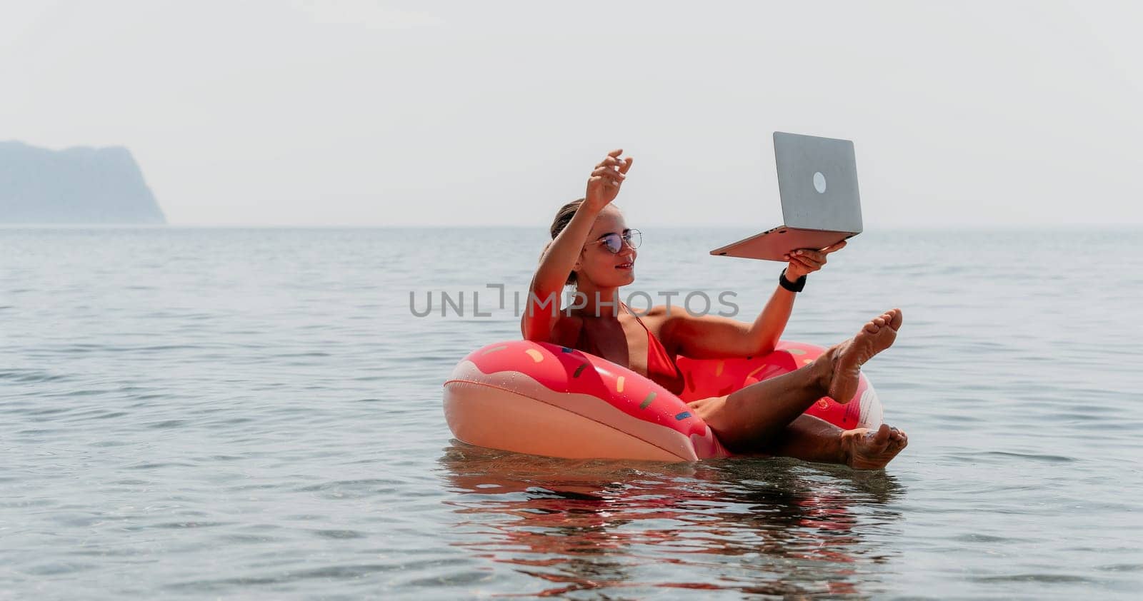 Woman freelancer works on laptop swimming in sea on pink inflatable ring. Happy tourist floating on inflatable donut and working on laptop computer in calm ocean. Freelance, remote working anywhere by panophotograph