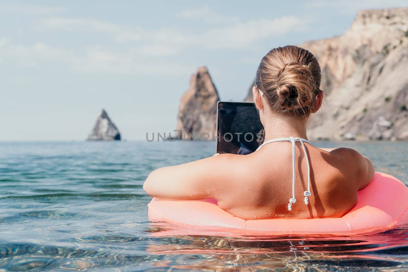 Woman freelancer works on laptop swimming in sea on pink inflatable ring. Happy tourist in sunglasses floating on inflatable donut and working on laptop computer in calm ocean. Remote working anywhere by panophotograph