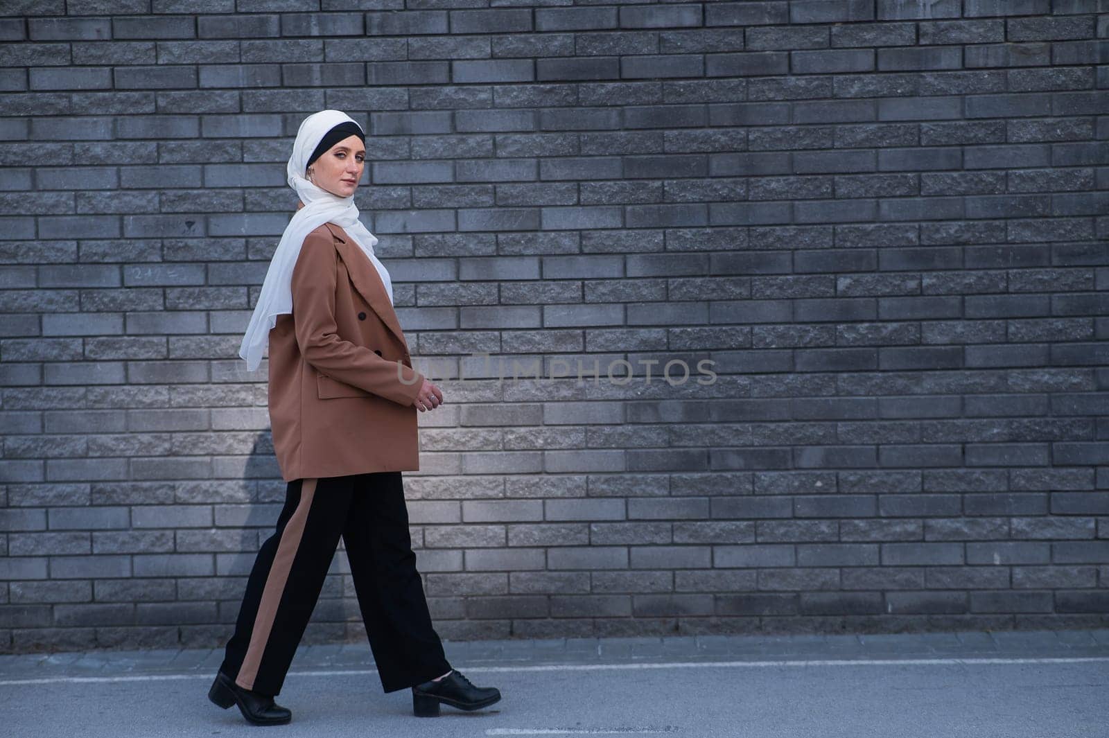 A young woman dressed in a hijab and a business suit walks along a brick wall. by mrwed54