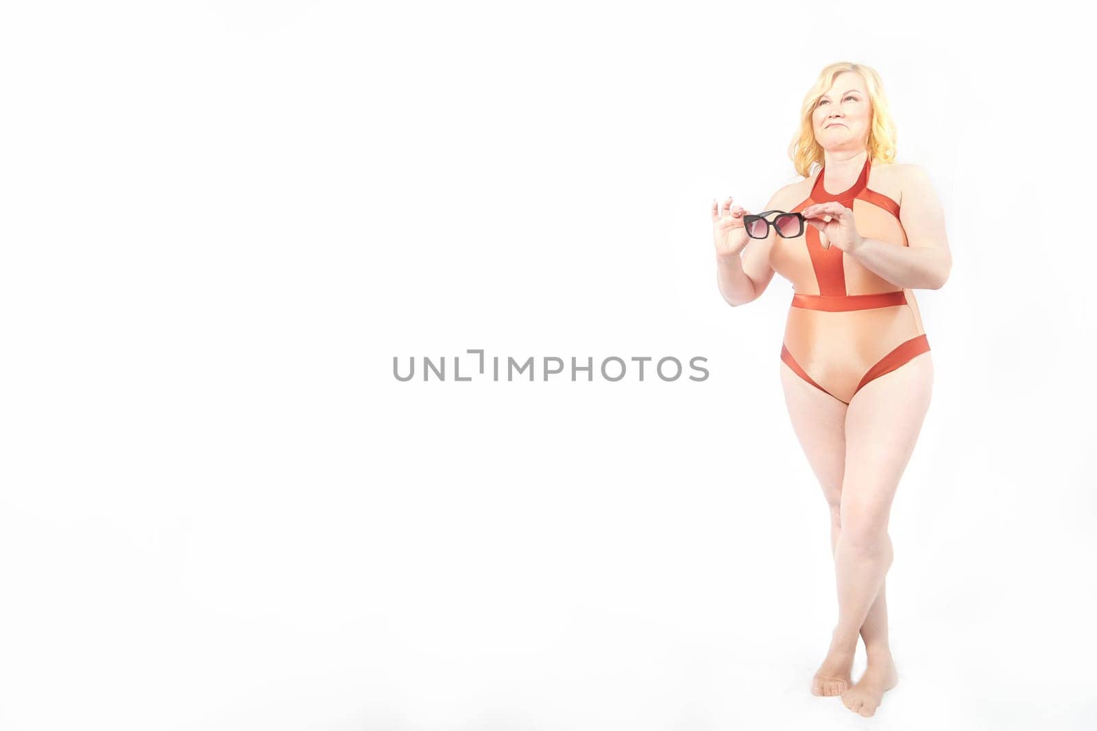 A cheerful, plump middle aged woman confidently poses in a red swimsuit or underwear and sunglasses, promoting the body positivity movement in a bright studio settings