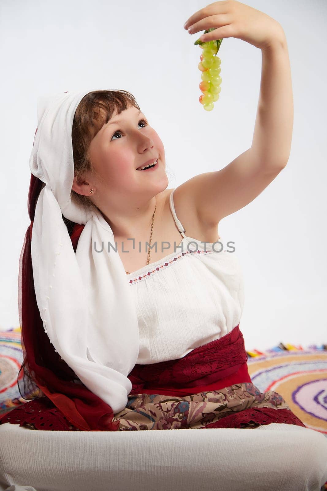 Portrait of Little girl in a stylized Tatar national costume with berries and a brush of grapes on a white background in the studio. Photo shoot of funny young teenager who is not professional model by keleny