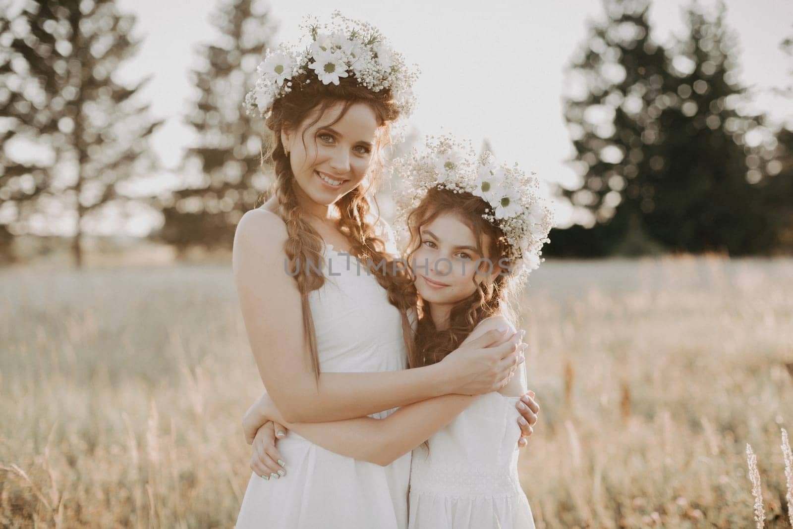 happy sisters in white dresses with floral wreaths and boho style braids in summer in a field in nature. Added a small grain simulation of film photography