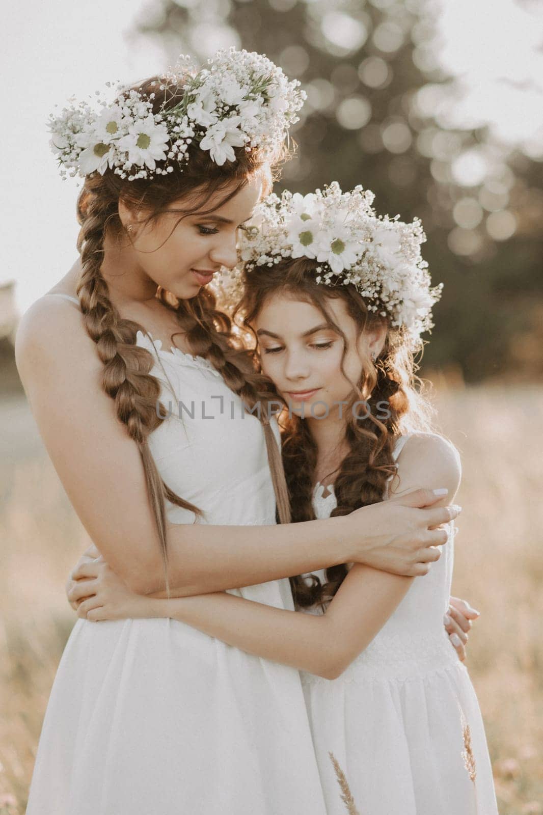 happy sisters in white dresses with floral wreaths and boho style braids in summer in a field in nature. Added a small grain simulation of film photography