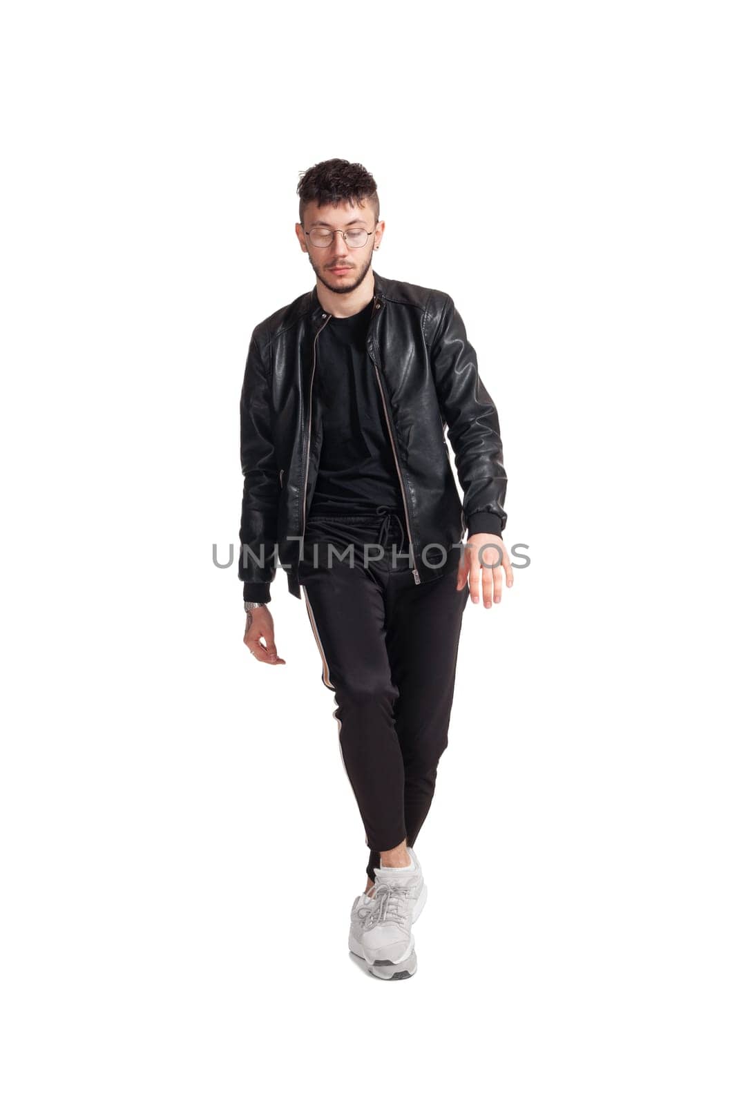 Full-length photo of a modern performer in glasses, black leather jacket, t-shirt, sports pants and light sneakers fooling around in studio. Indoor photo of a comical guy dancing isolated on white background. Music and imagination.