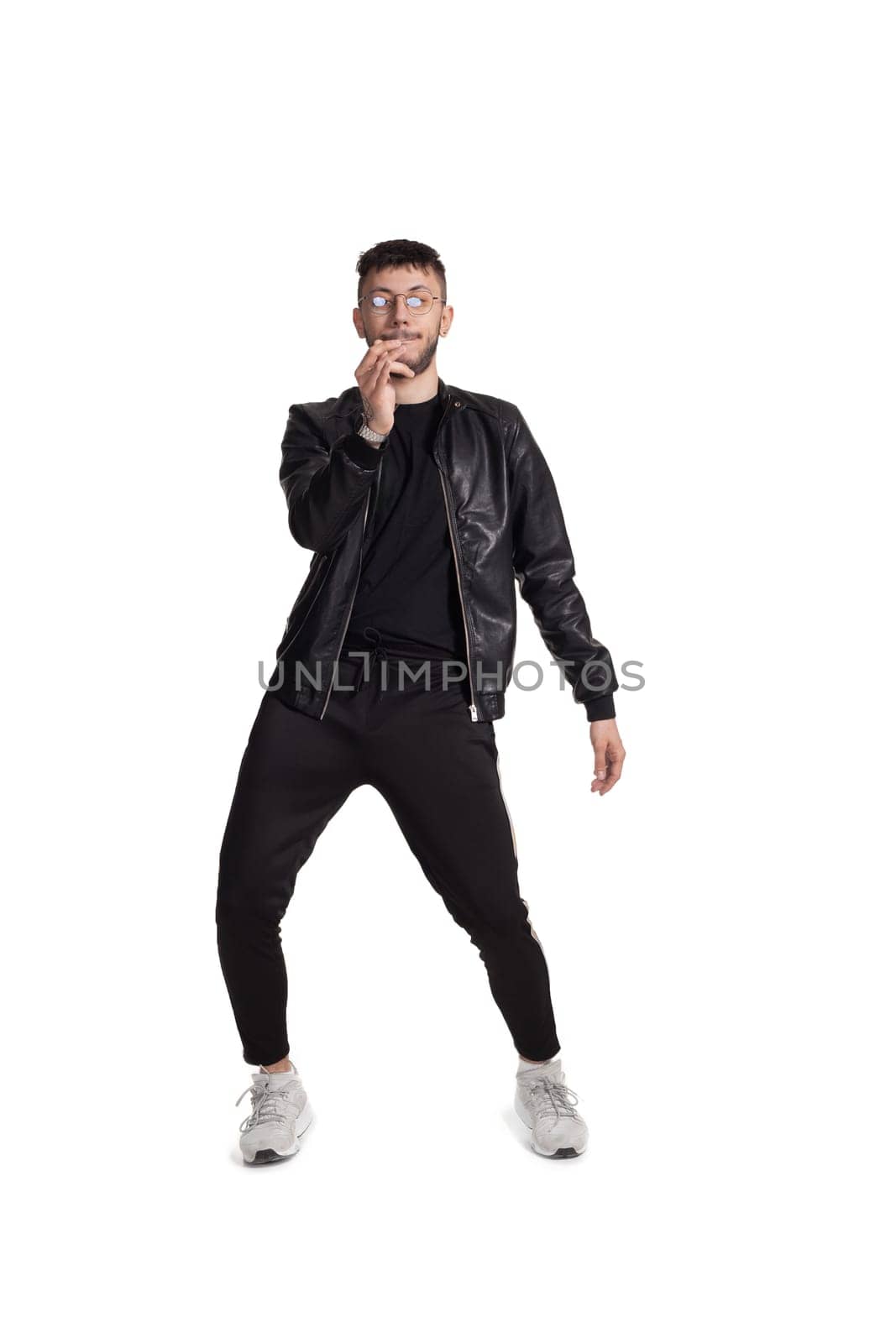 Full-length photo of a modern performer in glasses, black leather jacket, t-shirt, sports pants and light sneakers fooling around in studio. Indoor photo of a comical man dancing and gesticulating isolated on white background. Music and imagination.