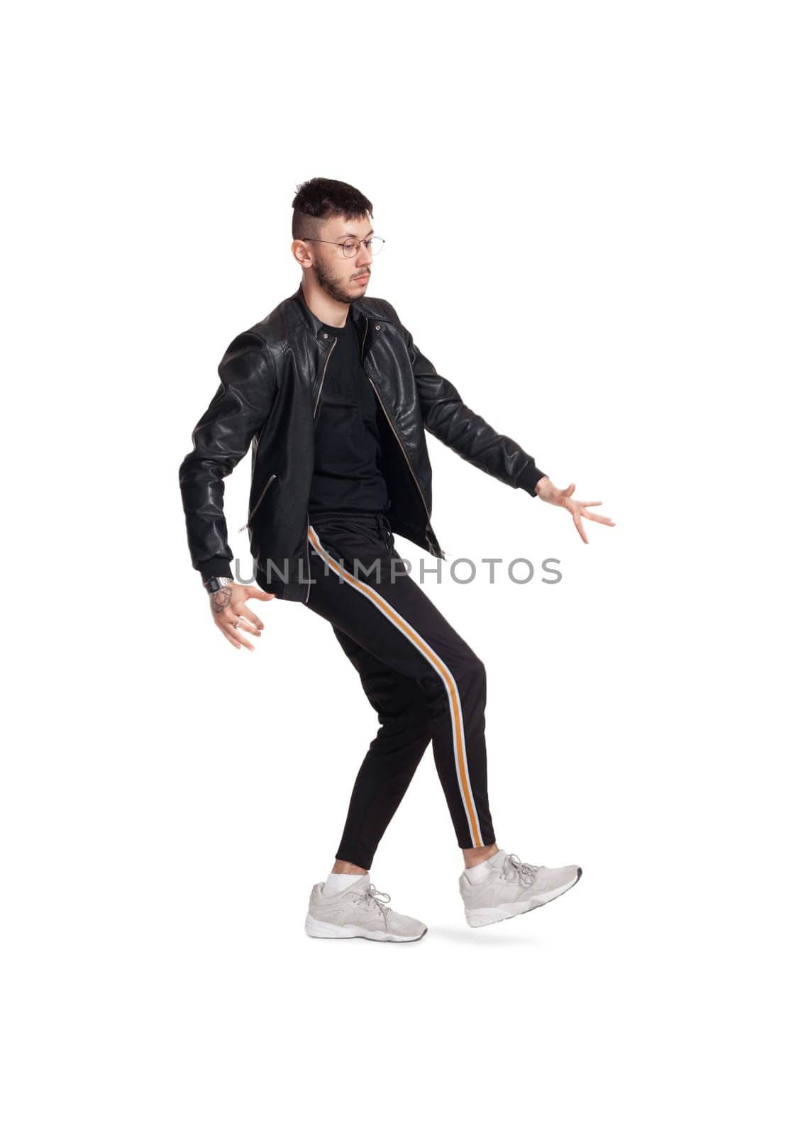 Full-length photo of a modern performer in glasses, black leather jacket, t-shirt, sports pants and light sneakers fooling around in studio. Indoor photo of a graceful man dancing sideways isolated on white background. Music and imagination.