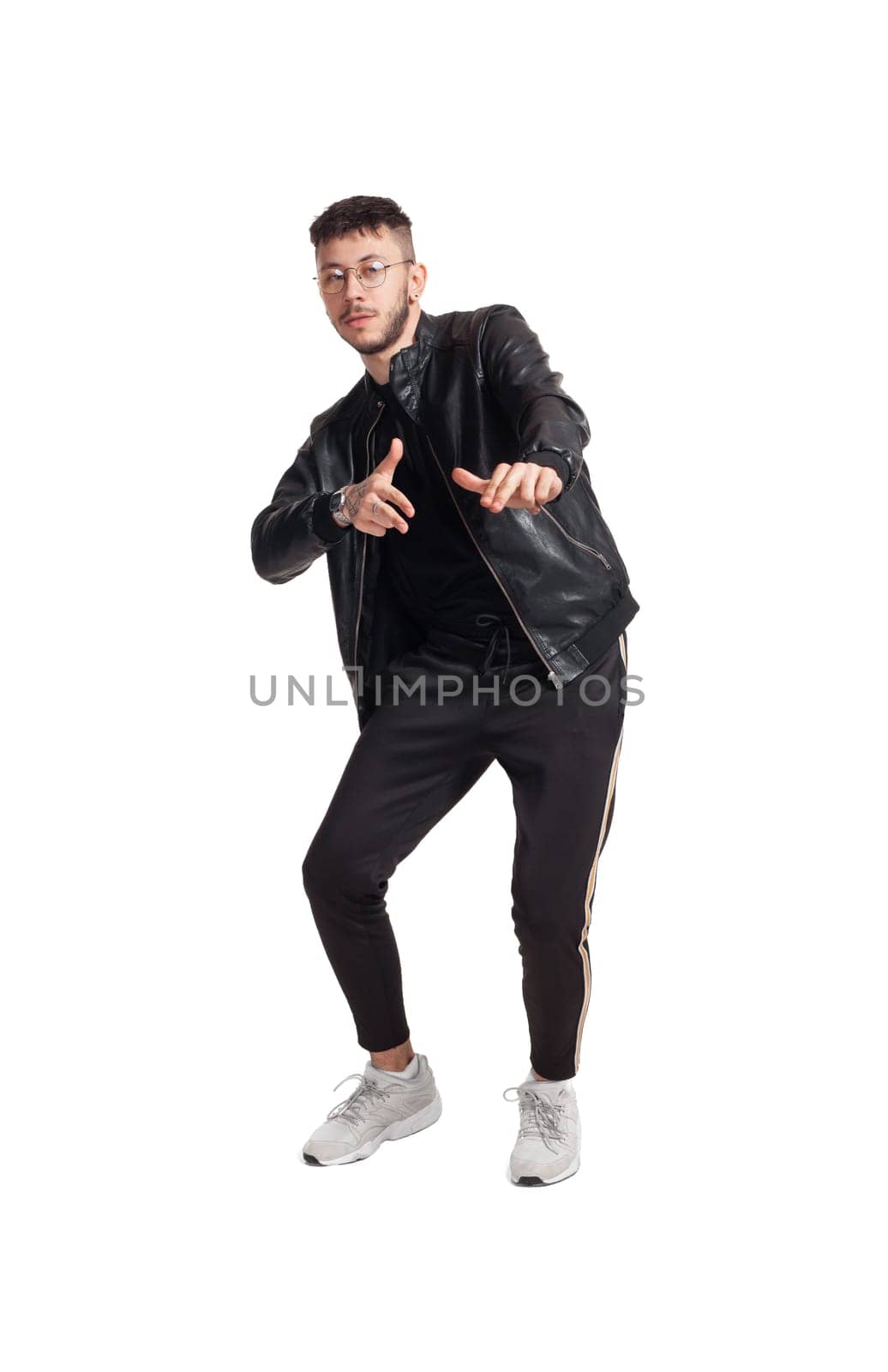 Full-length photo of a modern performer in glasses, black leather jacket, t-shirt, sports pants and light sneakers fooling around and gesticulating in studio. Indoor photo of a mischievous person dancing isolated on white background. Music and imagination.