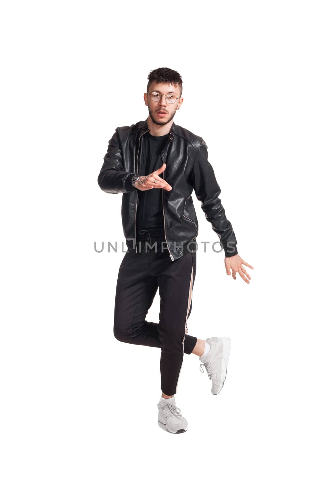 Full-length photo of a modern performer in glasses, black leather jacket, t-shirt, sports pants and light sneakers fooling around in studio. Indoor photo of a mischievous man dancing and looking at the camera isolated on white background. Music and imagination.