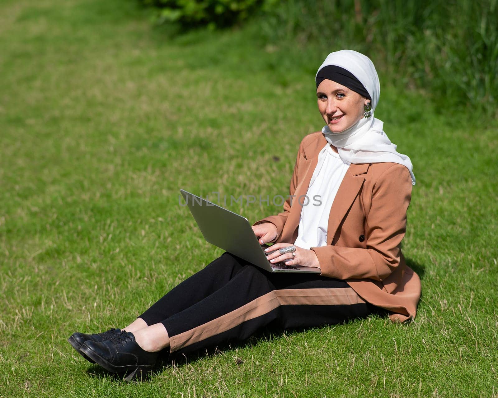 A young woman wearing a hijab sits on a lawn and uses a laptop outdoors. by mrwed54