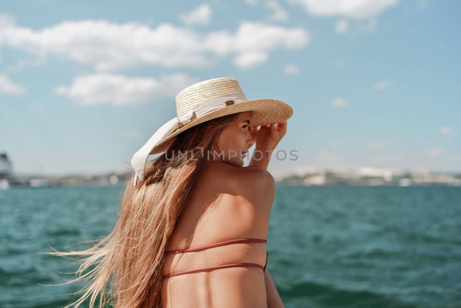 A woman in a swimsuit sits with her back holding a hat, looks at the ocean, sunny day, relaxes