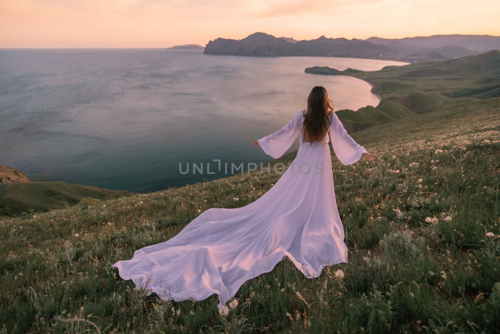 A woman in a long white dress stands on a hill overlooking the ocean. The scene is serene and peaceful, with the woman's dress flowing in the wind. The combination of the beautiful landscape