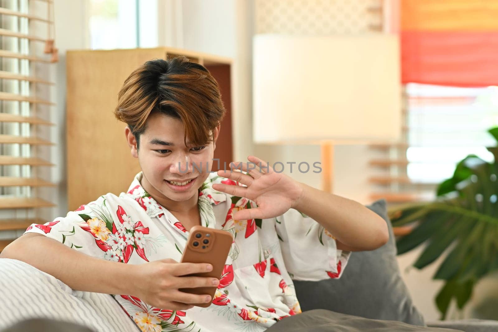 Cheerful young gay male sitting in living room and having video call on mobile phone with their friends.