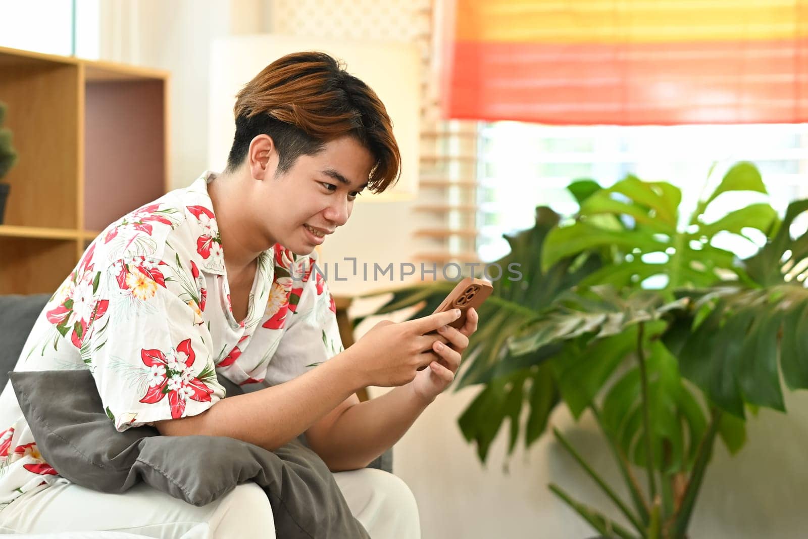 Happy young gay male reading social media post on smartphone, sitting on couch at home.