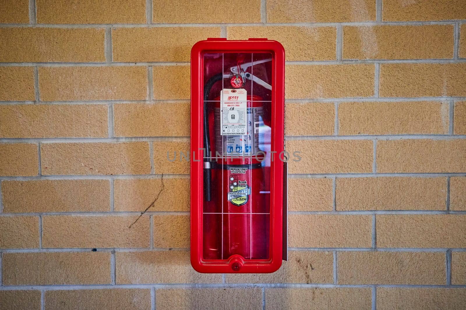 Vibrant red fire extinguisher in a glass case on a beige brick wall, Fort Wayne, emphasizing safety and readiness.