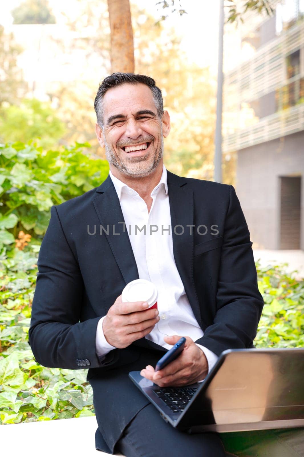 Satisfied with the results, the businessman walks down the street outside the office building, a mature boss holds a phone and coffee in his hands, writes messages and reads news online, using an app.