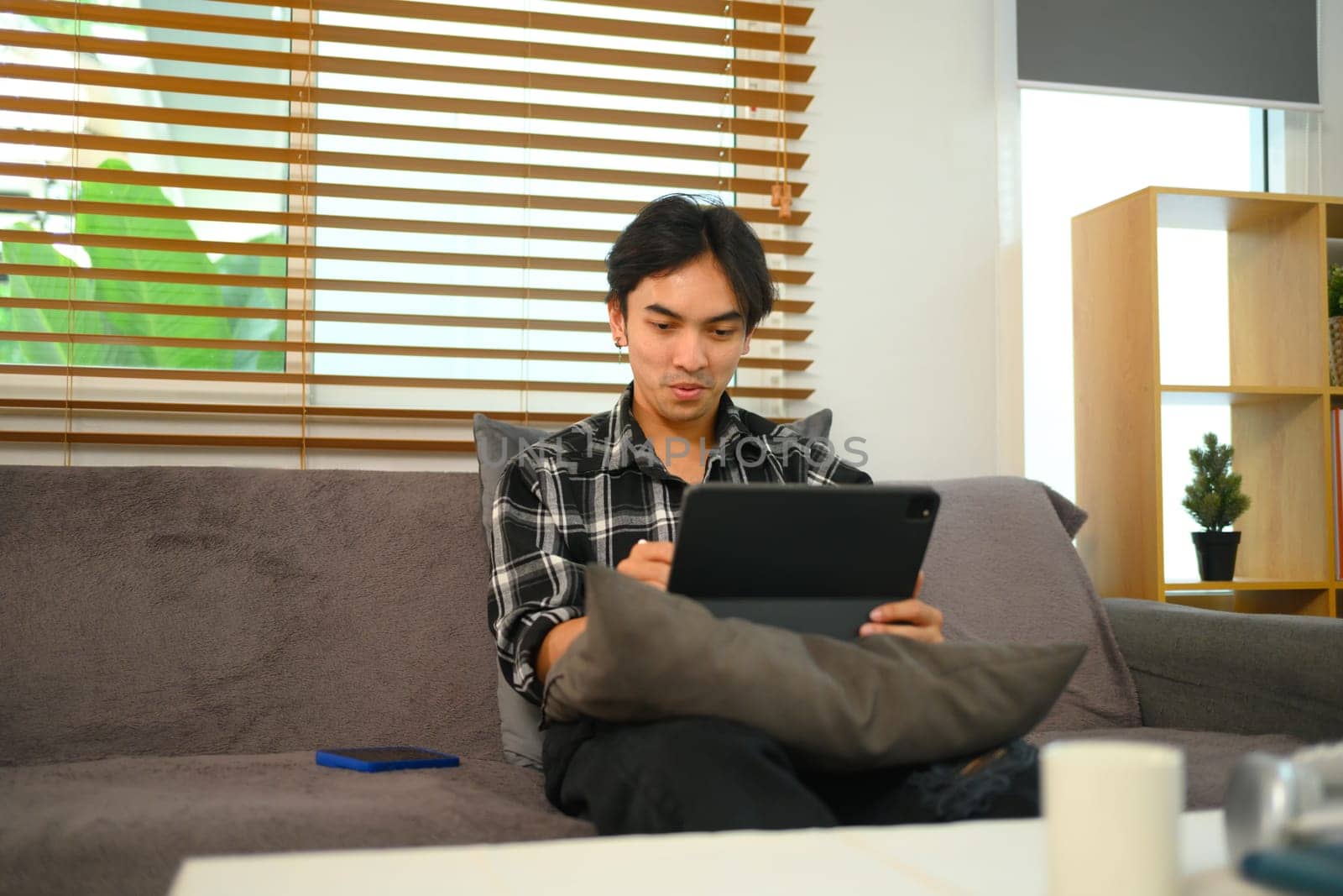 Smiling Young man watching video on digital tablet while relaxing on a couch at home by prathanchorruangsak