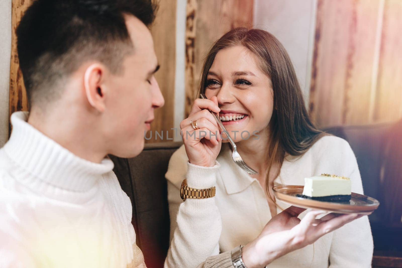 A cheerful young man, dressed in a white sweater, shares a delightful moment with his friend over a slice of cheesecake at a warm, inviting cafe. Their joyful interaction suggests a comfortable friendship as they savor the dessert together, surrounded by a wooden interior that adds to the cozy ambiance.