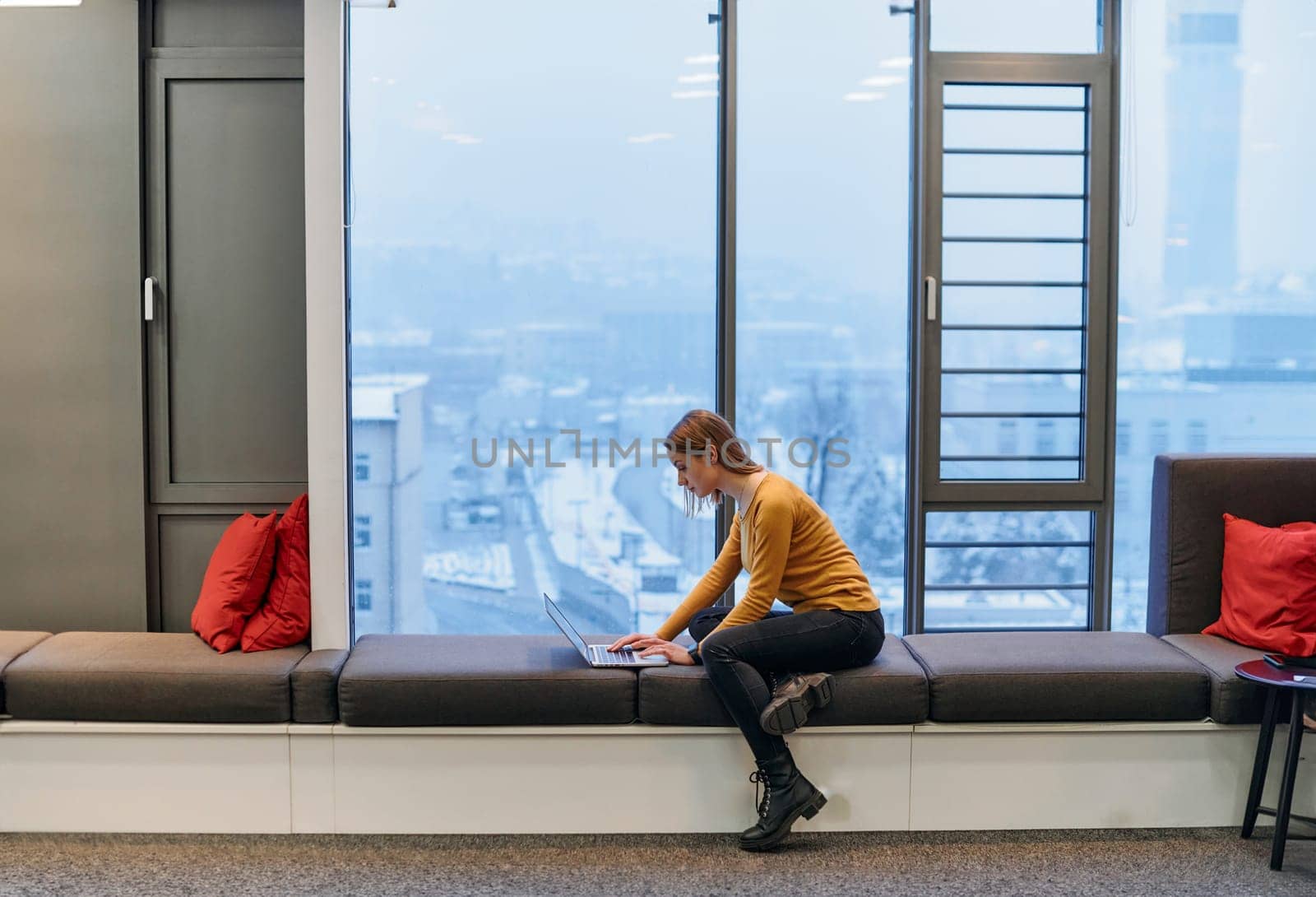 A businesswoman utilizes her laptop while seated by the window of a large corporate building, offering a picturesque view of the city skyline as her backdrop