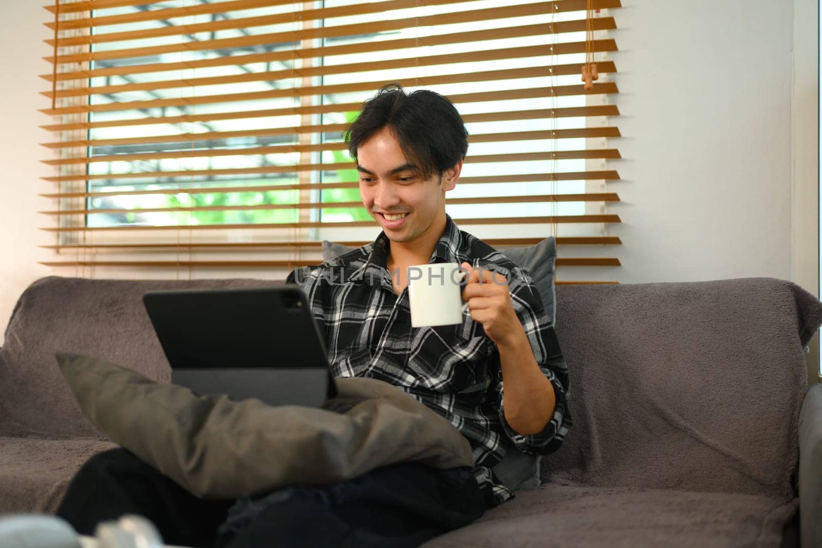 Cheerful asian man drinking coffee and using digital tablet for remote work at home by prathanchorruangsak