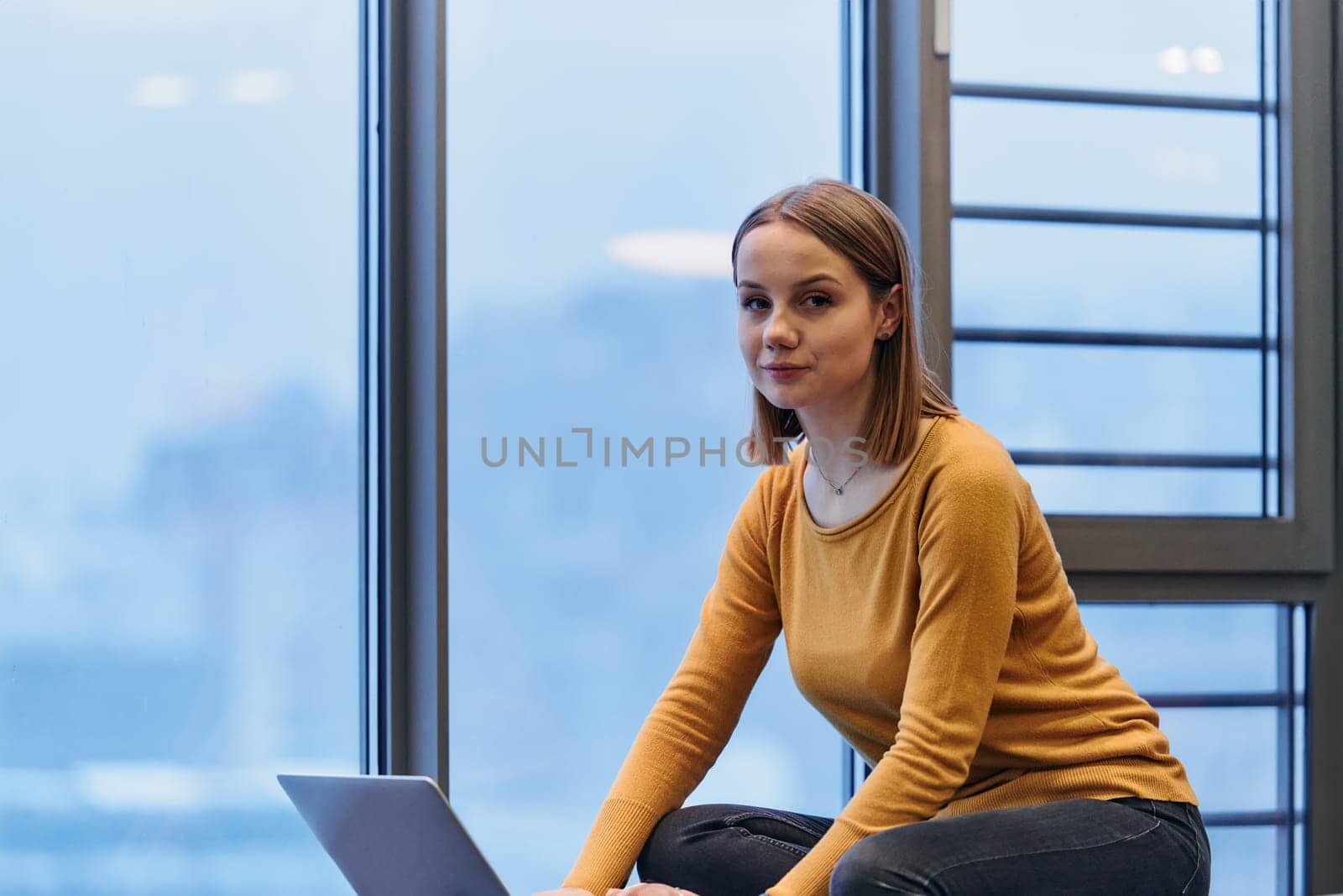 A businesswoman utilizes her laptop while seated by the window of a large corporate building, offering a picturesque view of the city skyline as her backdrop