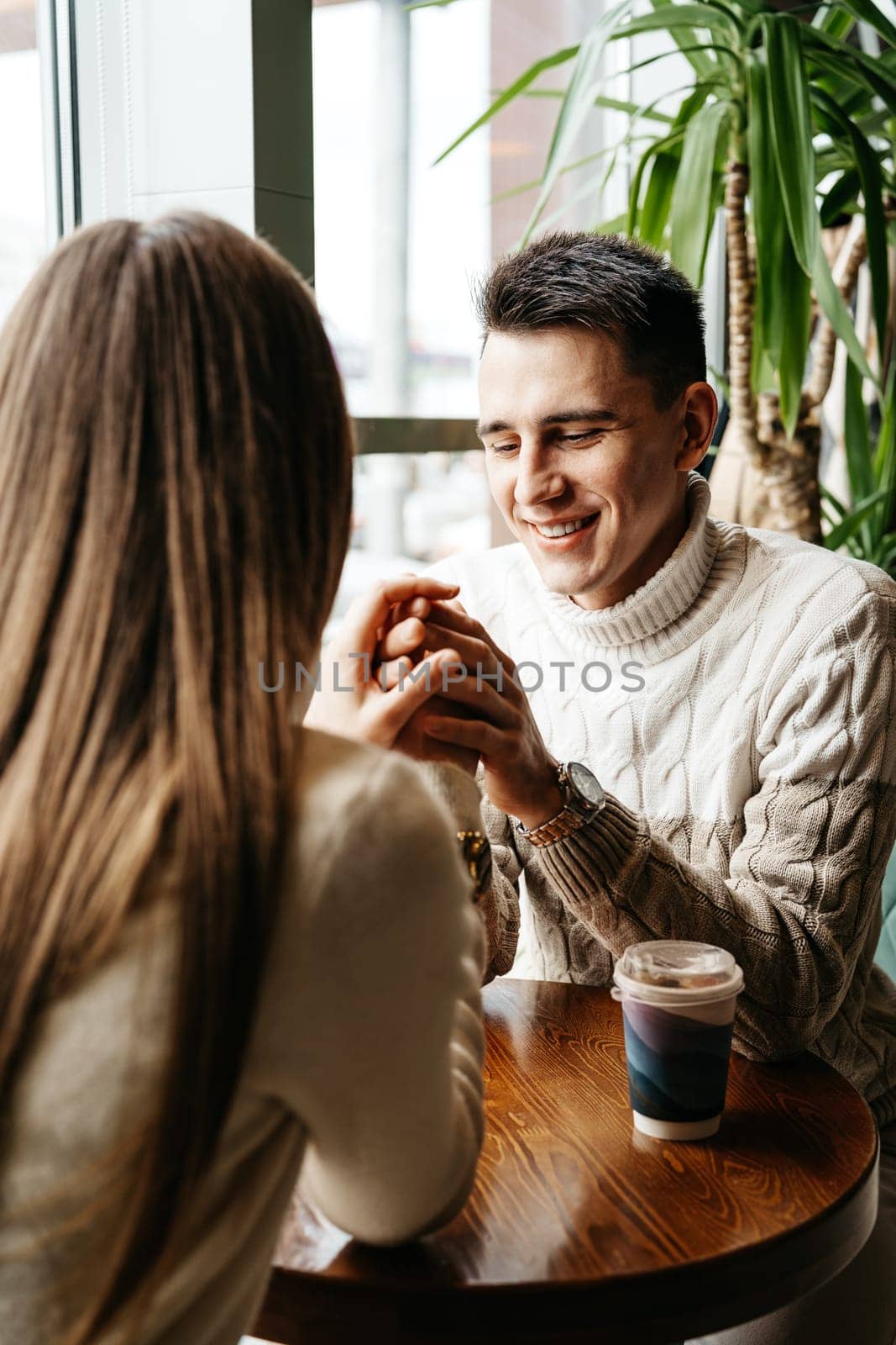 A cheerful man in a knit sweater is enjoying a chat with a woman at a wooden table in a quaint cafe. A drink in a disposable cup suggests a relaxed, informal setting, and natural light spilling from the window adds warmth to the scene. Their engagement in conversation captures a moment of genuine connection and companionship.