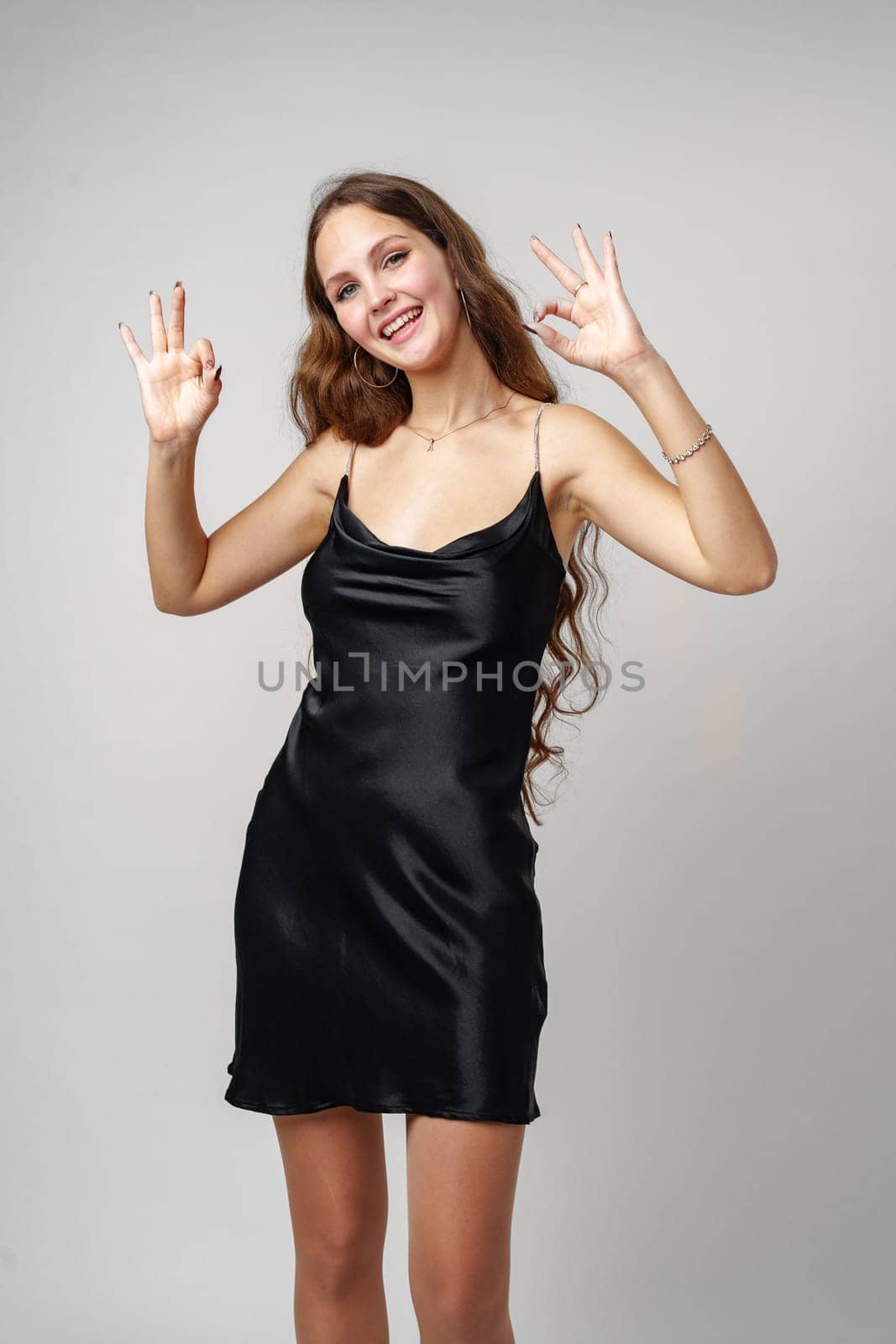 A cheerful young woman with long brown hair is seen giving an okay gesture with both hands, wearing a sleek black dress that suggests a formal or festive occasion. Her joyous expression and the simplicity of the background draw attention to her gesture and attire.