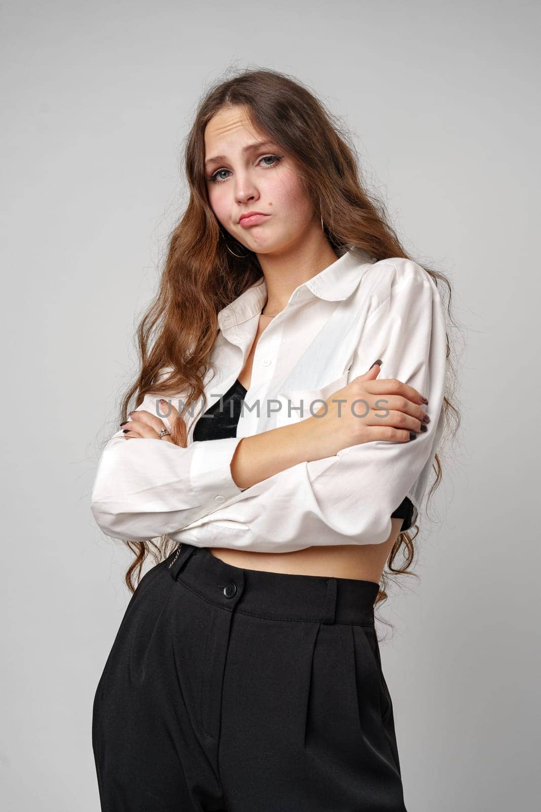 A young woman with long, wavy brown hair and light makeup exhibits a playful yet melancholic pout. She is wearing a casual white button-up shirt, with the top buttons undone, giving the impression of a relaxed or end-of-day mood. The backdrop is plain and grey, drawing full focus to her expressive face.