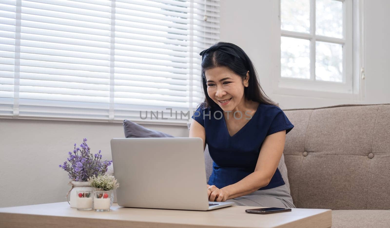 Happy Asian retired woman in her 60's enjoying social media on laptop while relaxing in living room..
