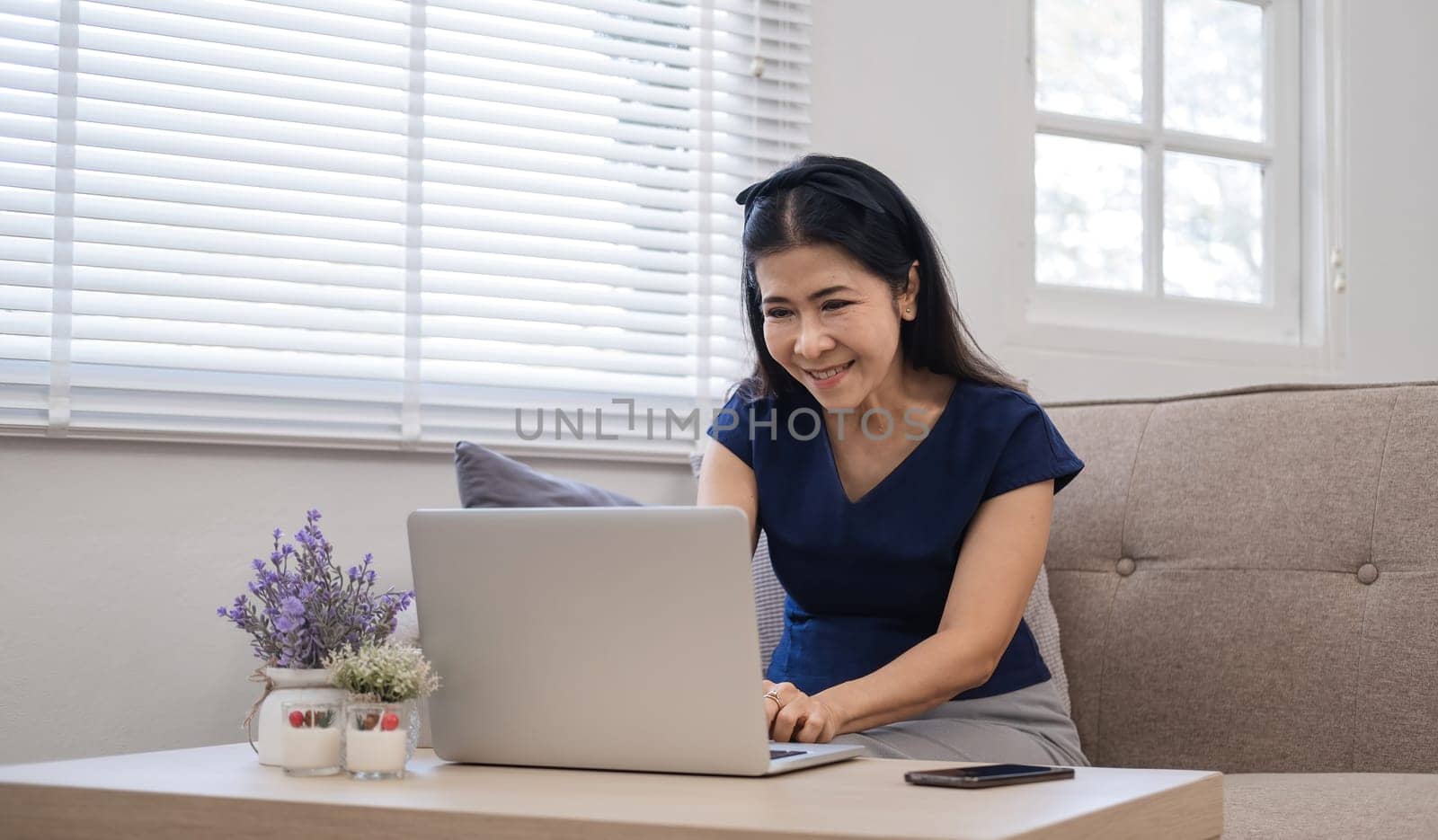 Happy Asian retired woman in her 60's enjoying social media on laptop while relaxing in living room. by wichayada