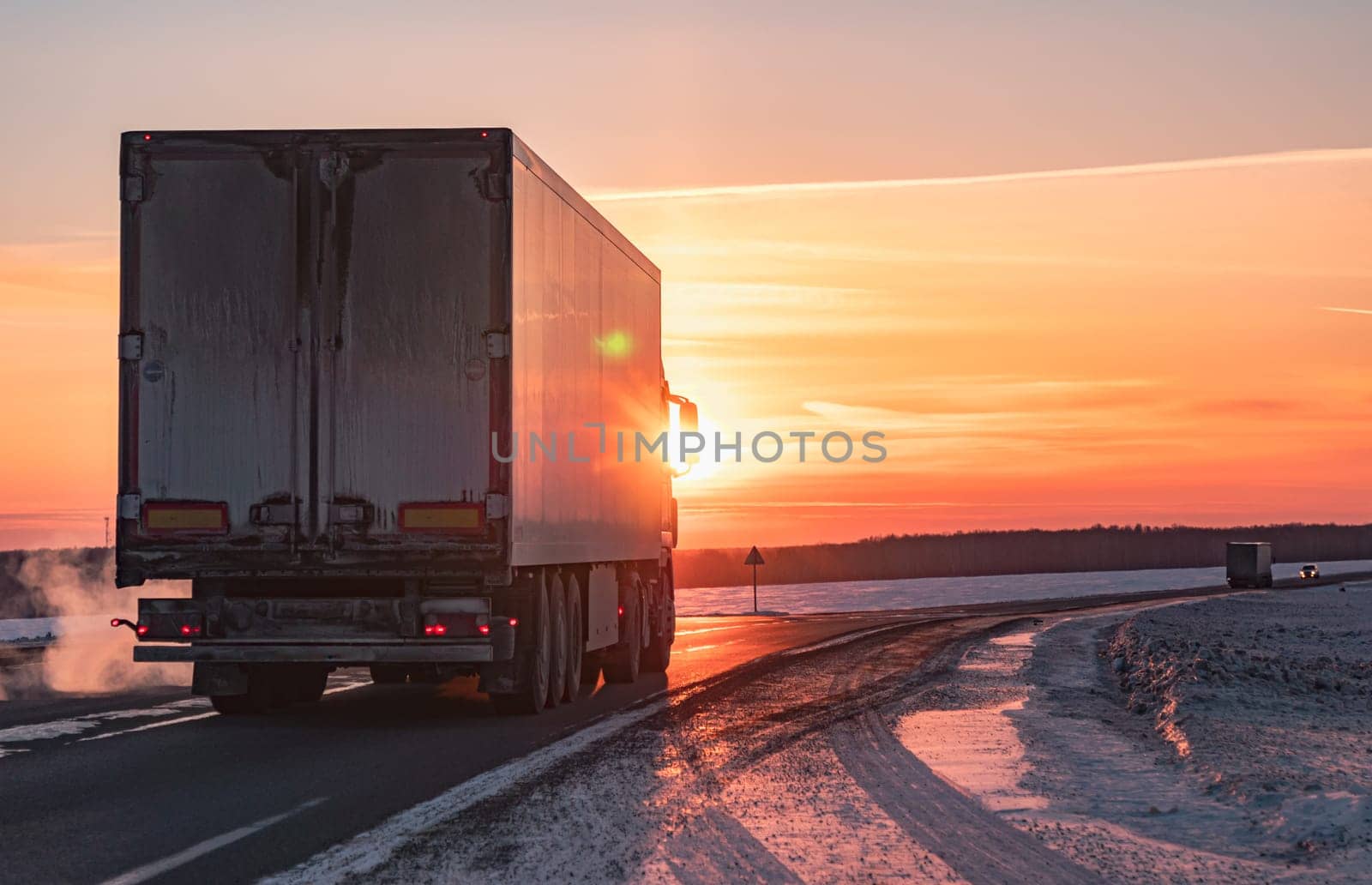 A semi truck cruises down a wintry highway as the sun sets on the horizon, casting a warm glow over the icy road and surrounding landscape.