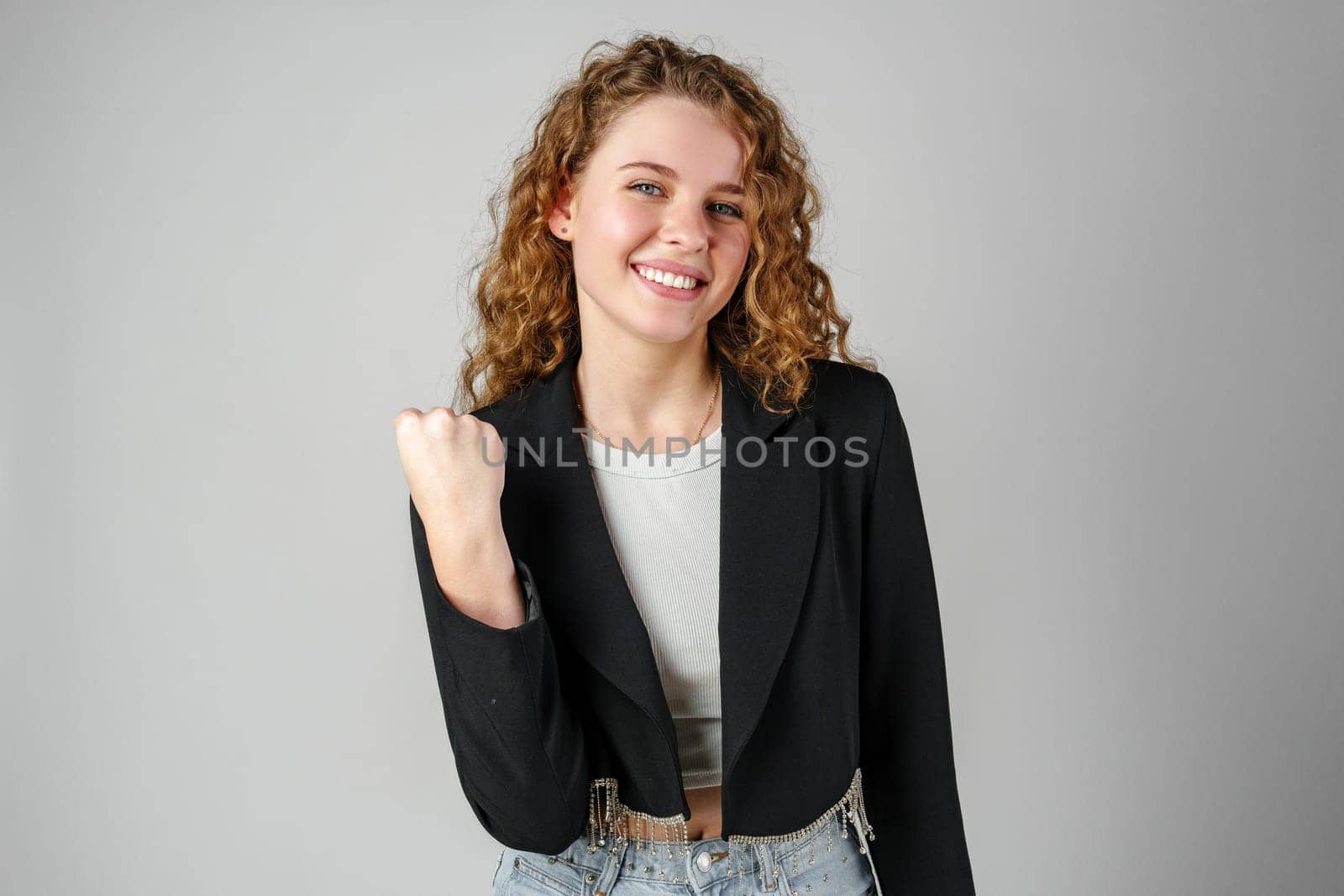 Excited Young Woman Celebrates Success With Raised Fists Against a Gray Background by Fabrikasimf