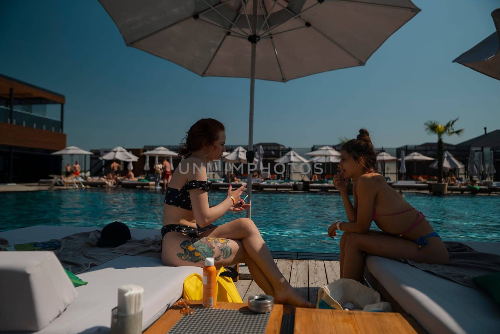 Two young women in bathing suits sit together by the pool, chatting and laughing. by teksomolika