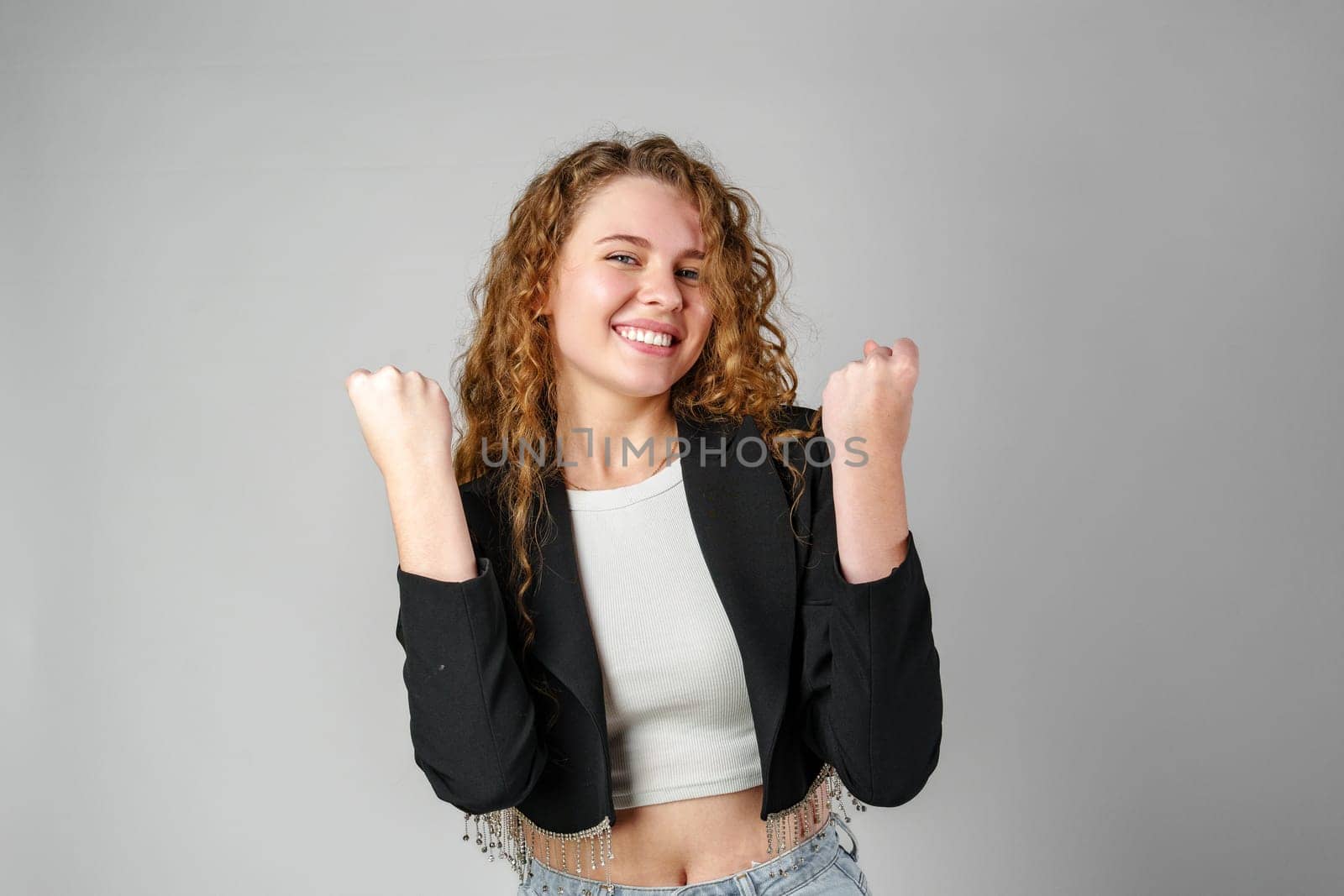 Excited Young Woman Celebrates Success With Raised Fists Against a Gray Background by Fabrikasimf