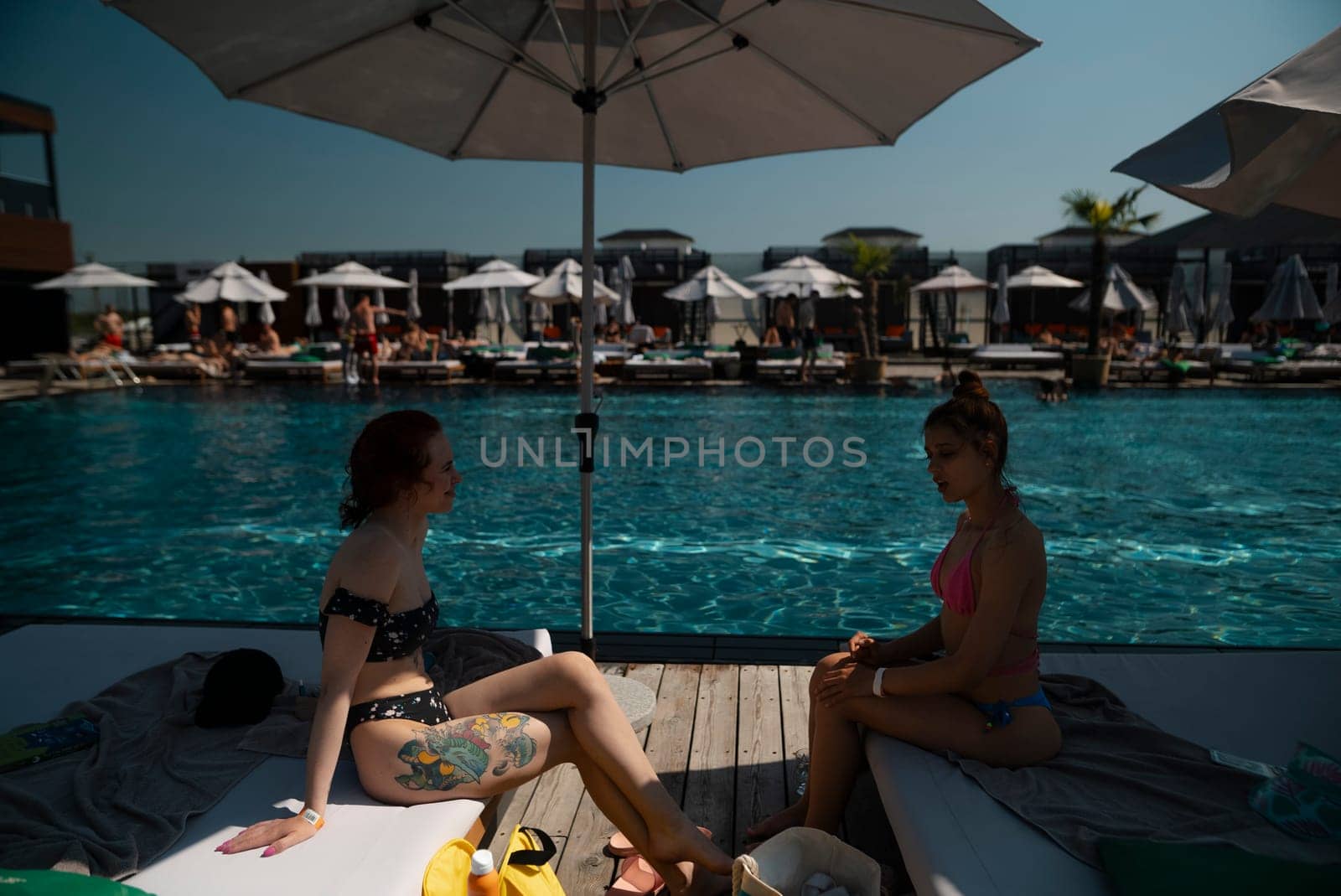 A couple of young ladies in swimsuits sit by the pool, savoring the moment. by teksomolika