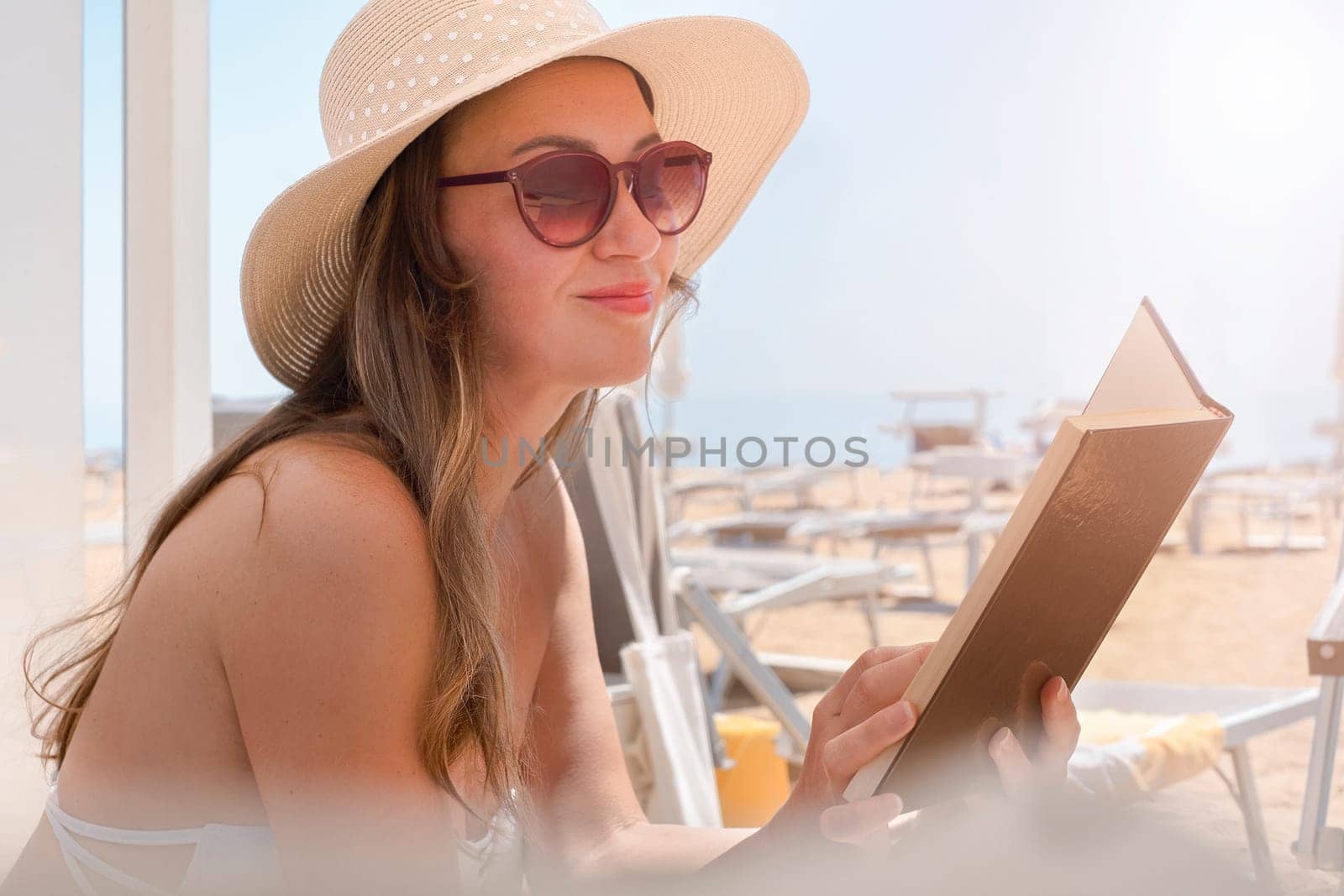 Portrait of a young woman with straw hat, relaxing on the beach, reading a book. Summer concept