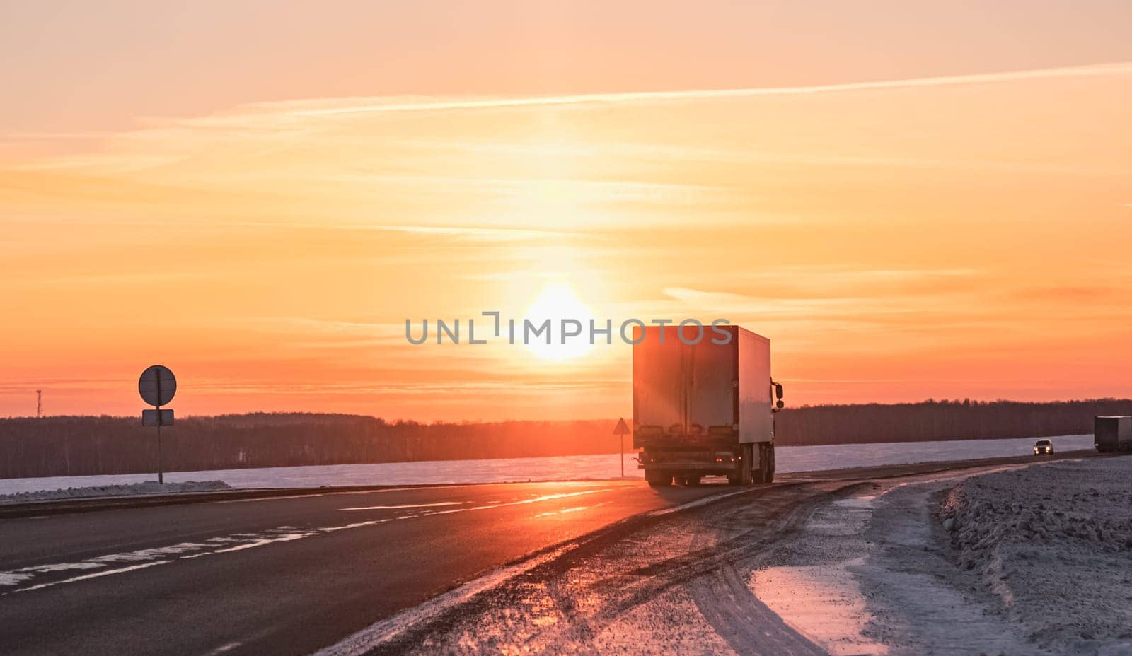 A semi truck cruises down a wintry highway as the sun sets on the horizon, casting a warm glow over the icy road and surrounding landscape.