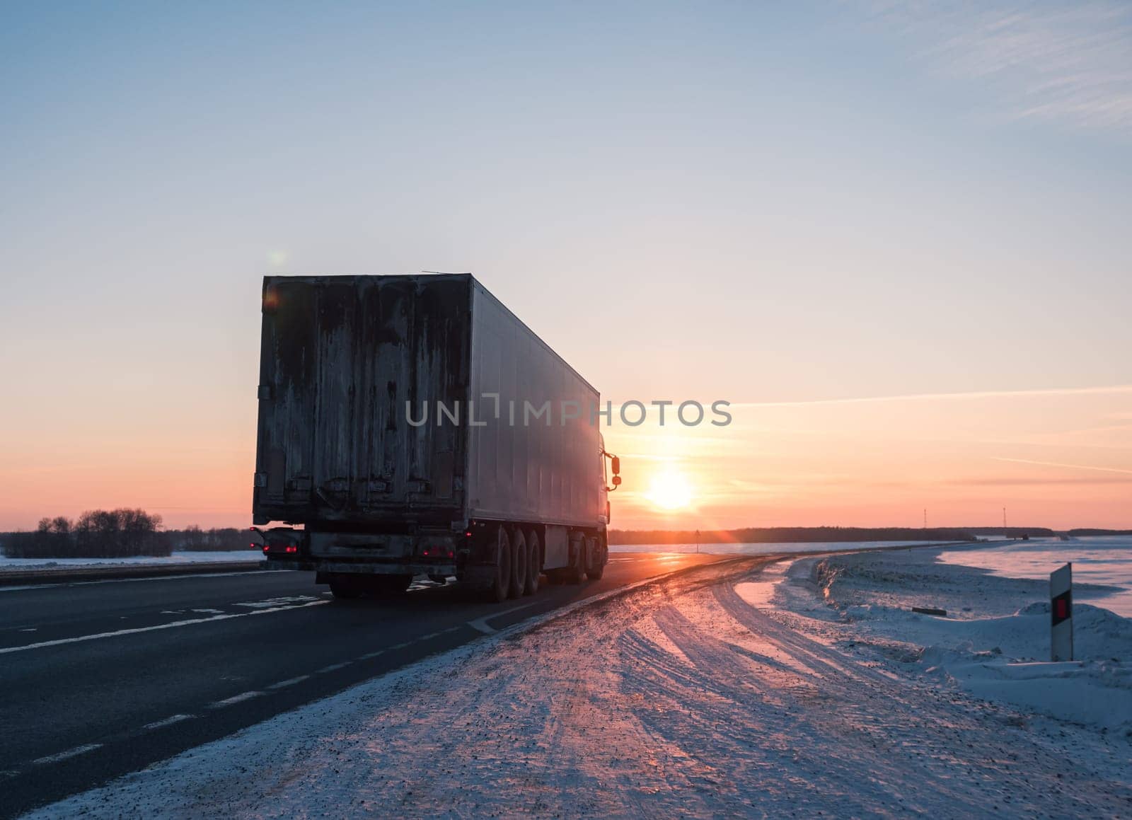 A semi truck cruises down a wintry highway as the sun sets on the horizon, casting a warm glow over the icy road and surrounding landscape.