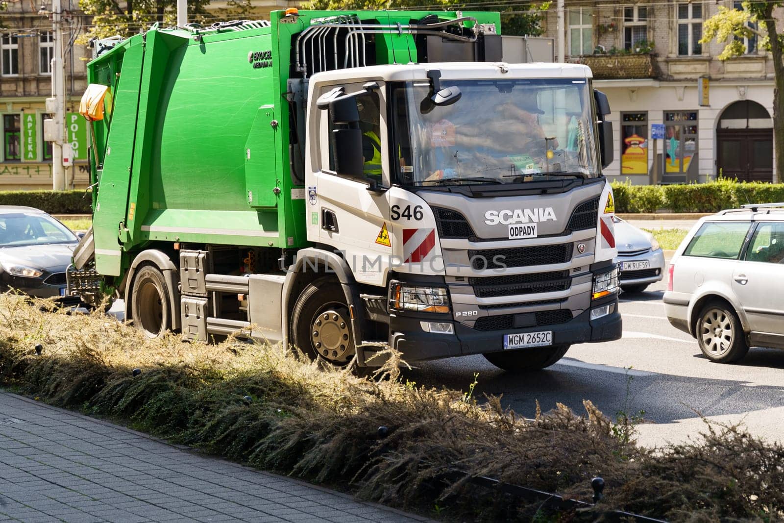 Wroclaw, Poland - August 4, 2023: A green and white Scania garbage truck is actively collecting waste along a busy city street with buildings and other vehicles visible in the background.
