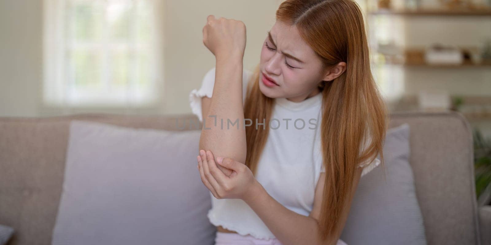 Young woman feeling elbow pain while sitting on sofa at home by nateemee