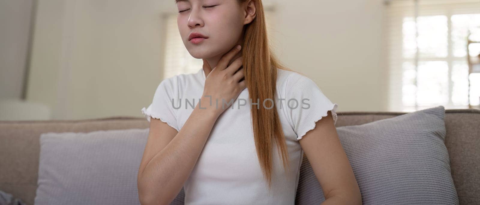 Young woman experiencing throat pain while sitting on a sofa at home by nateemee