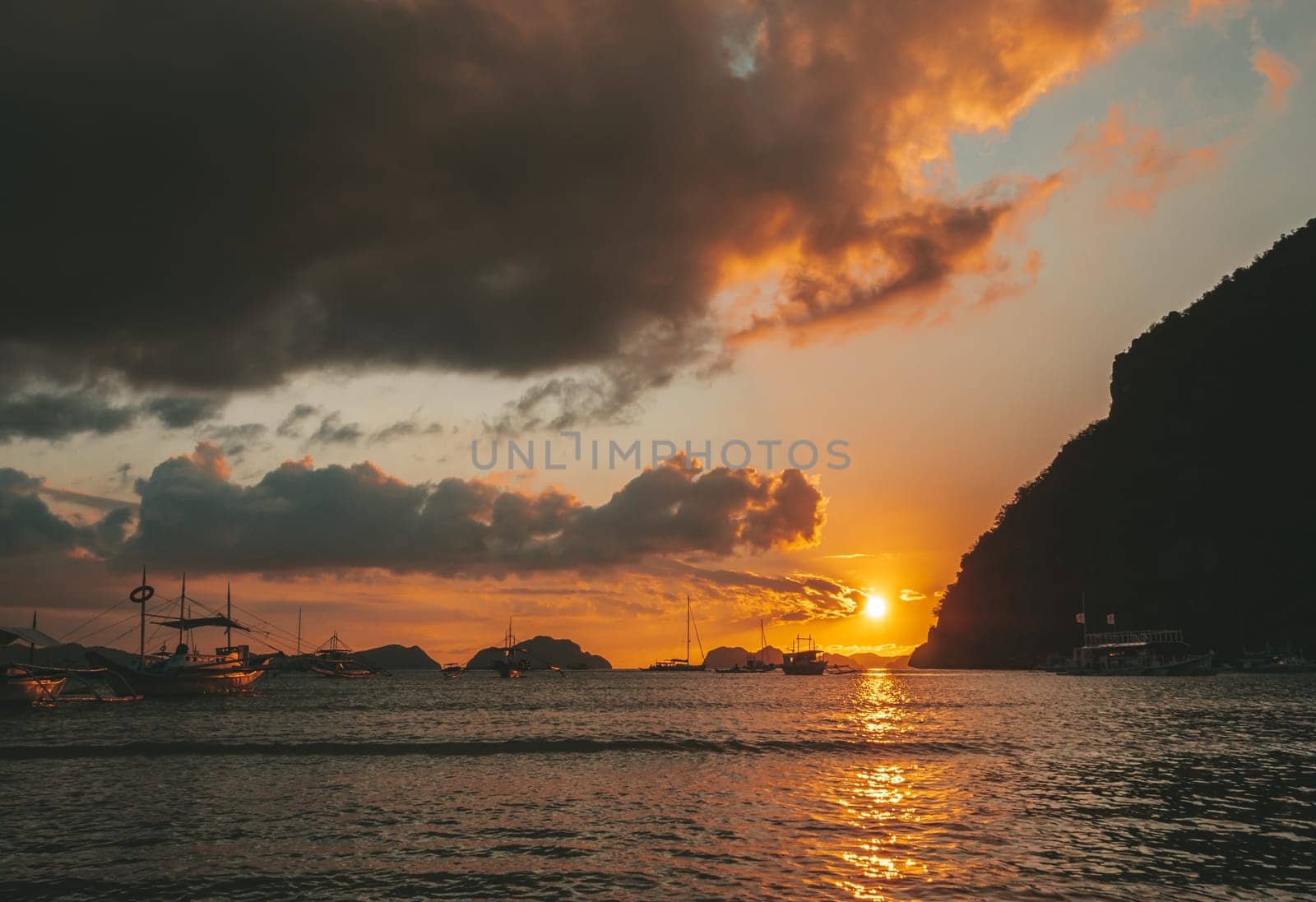 Multiple boats are anchored in a calm bay under a vibrant sunset. The sky glows with shades of orange and gold, while dark clouds create dramatic contrasts.